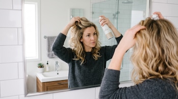 Una mujer sostiene un frasco de champú en seco mientras lo aplica en las raíces de su cabello frente al espejo de un baño.
