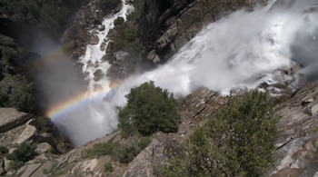 Vista aérea de una potente cascada blanca cayendo por un acantilado rocoso gris, con un arcoíris en la niebla y árboles verdes en primer plano