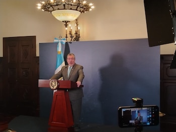 Hombre en traje en podio de madera con emblema. Fondo con bandera de Argentina y panel azul. Lámpara de techo ornamental. Teléfono móvil grabando