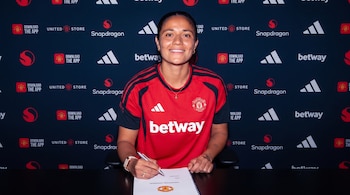 Una mujer con camiseta deportiva roja y negra sonríe mientras firma un documento sobre una mesa frente a una pared con logotipos de marcas.