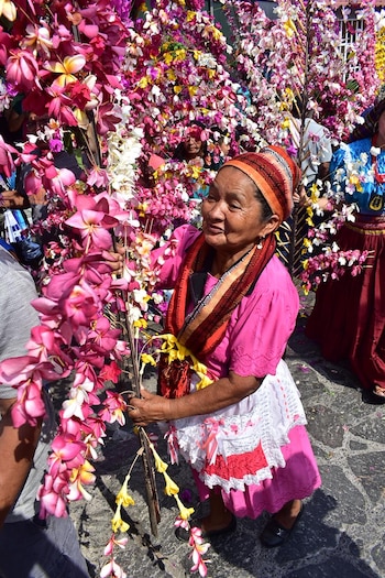 Una mujer sonriente con turbante y blusa rosa sostiene ramas con flores artificiales rosadas, blancas y amarillas, rodeada de más flores