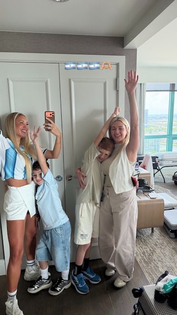 Dos mujeres y dos niños sonríen y posan en una habitación. Una mujer viste una camiseta de la selección argentina. Tres banderas argentinas decoran una puerta