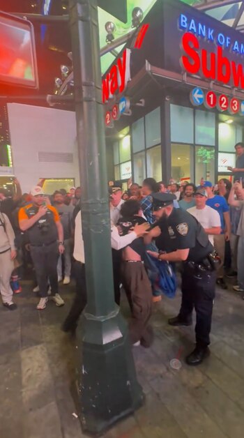 Un oficial de policía interactúa con personas entre una multitud en una calle urbana por la noche, con carteles de "Bank of America" y "Subway" visibles
