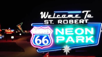 Un letrero de neón azul y blanco "Welcome To St. Robert NEON PARK" con un escudo azul de la Ruta 66, iluminado de noche sobre un fondo oscuro