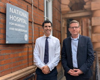 Dos hombres con ropa formal posan junto a una placa con la inscripción National Hospital for Neurology and Neurosurgery en una pared de ladrillos