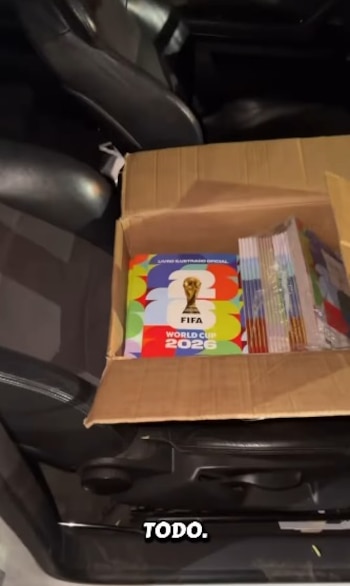 Una caja de cartón abierta en el asiento de un coche, llena de álbumes coloridos y paquetes de figuritas del Mundial 2026 de la FIFA