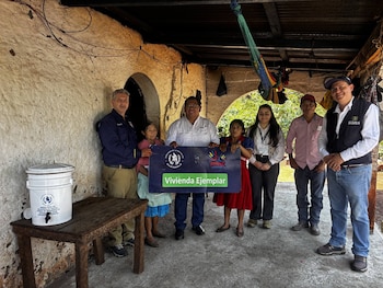 Varias personas, incluyendo hombres, mujeres y niños, sentadas en mesas largas comiendo de platos con comida variada y bebidas en vasos de plástico