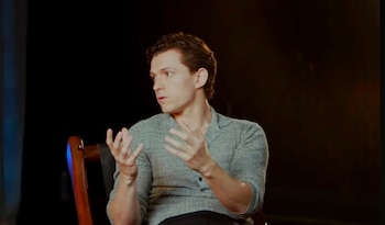 El actor Tom Holland, de perfil, sentado y gesticulando con las manos. Viste una camisa gris y su cabello castaño está peinado hacia atrás. Fondo oscuro