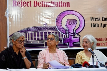 Tres mujeres indias sentadas en una mesa con micrófonos. Detrás, un cartel beige con el texto "Reject Delimitation", el Parlamento indio y un símbolo femenino con puño