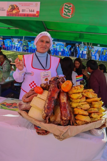Mujer con gorro blanco y delantal exhibe un gran plato de Chiriuchu con cuyes, gallina, salchichas, quesos y tortillas, en un puesto de mercado al aire libre