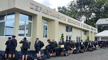 Edificio beige, letrero azul con "DEPARTAMENTO EDUCATIVO Y DOCTRINA SGD-PNC", grupo de personas con uniformes oscuros y equipaje, árboles al fondo, cielo nublado