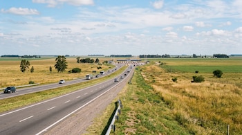 Una autopista con automóviles y camiones atraviesa un área rural con vegetación y cabinas de peaje al fondo bajo un cielo azul