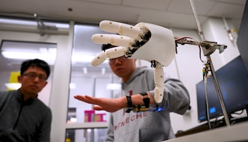 Una mano robótica blanca en primer plano, con un hombre que lleva una muñequera de tecnología en su mano derecha y otro hombre borroso al fondo en un laboratorio