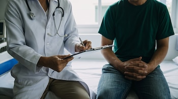 Un médico con bata blanca y estetoscopio escribe en un portapapeles junto a un paciente sentado con camiseta verde oscuro y jeans, en una cama de hospital.