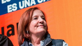 Alejandra Vigo con camisa clara y chaleco oscuro de abrigo, con las manos entrelazadas delante de una pantalla con texto