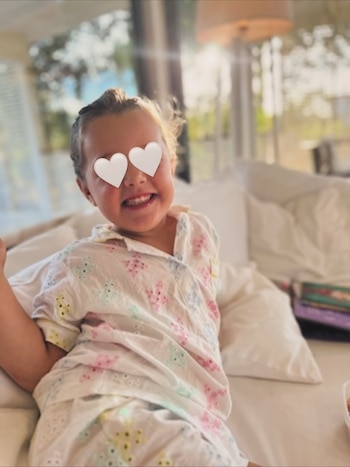 Una niña pequeña sonriendo, con sus ojos cubiertos por corazones blancos, sentada en una cama con sábanas blancas, vistiendo un pijama blanco con flores de colores