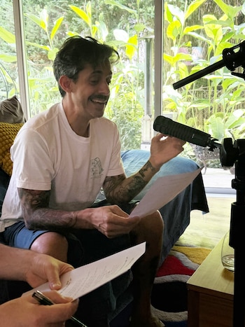 Hombre de bigote con camiseta blanca y brazos tatuados, sentado y sonriente, sosteniendo un micrófono negro y papeles. Se ve una ventana con vegetación