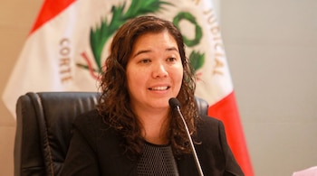 Una mujer de cabello castaño habla frente a un micrófono con una bandera peruana detrás