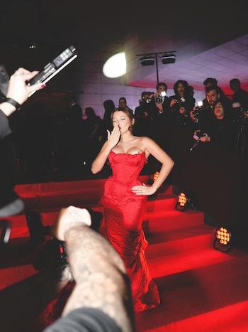 Una mujer con un vestido rojo sin tirantes y corsé posa en una alfombra roja, enviando un beso. Luces brillantes y fotógrafos con cámaras se ven al fondo