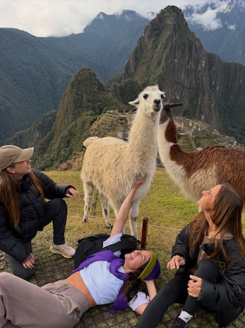 Tres mujeres adultas junto a dos llamas en un paisaje montañoso con ruinas de piedra y cielo nublado