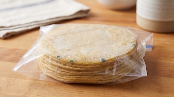 Una pila de tortillas dentro de una bolsa de plástico transparente con gotas de condensación y pequeñas manchas verdes sobre una mesa de madera.