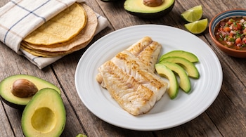Un plato blanco con dos filetes de pescado y rodajas de palta. Varias tortillas, dos mitades de palta, gajos de lima y un tazón de salsa en una mesa de madera.