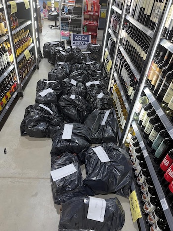 Múltiples bolsas de basura negras apiladas en el suelo de un pasillo, rodeadas por estanterías llenas de botellas de bebidas y un cartel de Aduana