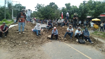 Un grupo de personas, en su mayoría hombres, se encuentra sentado y de pie sobre un montículo de tierra y rocas que bloquea una carretera asfaltada