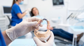 Manos con guantes sujetan una prótesis dental removible y un modelo de mandíbula; al fondo, un profesional con mascarilla y un paciente en una silla dental.
