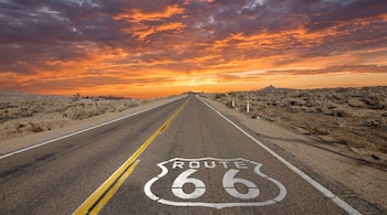 Carretera de la Ruta 66 con un escudo blanco pintado en el asfalto, extendiéndose hacia el horizonte bajo un cielo al atardecer de tonos naranja y morado sobre un desierto.