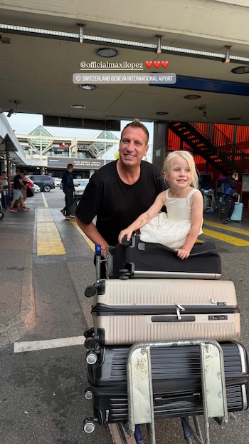 Maxi López junto a una niña rubia sentada sobre varias maletas apiladas en un carro de equipaje, en la entrada de un aeropuerto