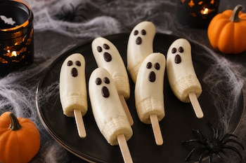 Seis trozos de plátano en brochetas cubiertos con crema blanca en un plato negro, rodeados de calabazas pequeñas y telarañas decorativas.