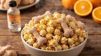Un tazón con palomitas de maíz, ralladura de naranja y piezas de pan con forma de hueso, junto a una botella de esencia y naranjas sobre madera.
