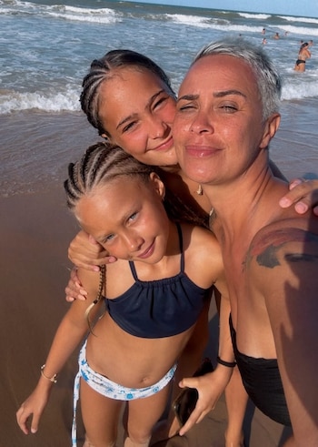 Primer plano de Josefina Pouso, con cabello gris corto, abrazando a sus dos hijas en la playa. La hija mayor sonríe detrás, la menor mira a cámara. Fondo de mar con olas