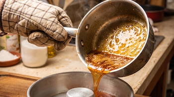 Una mano con guante de cocina sostiene una sartén con caramelo líquido y lo vierte en un molde para flan metálico sobre una superficie de madera.