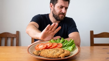 Un hombre con barba en una mesa hace un gesto de alto hacia un plato con una milanesa, lechuga y rodajas de tomate.