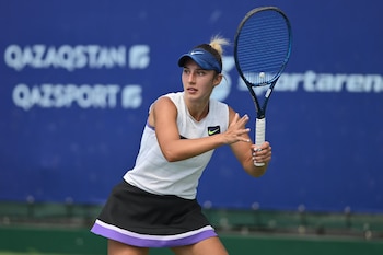 Anastasia Sukhotina, tenista, con visor azul, top blanco y falda negra, sujeta una raqueta en una cancha con fondo azul y texto borroso