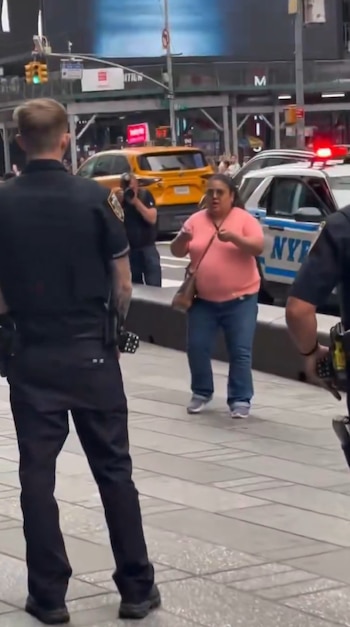 Una mujer con camiseta rosa sostiene objetos en sus manos frente a un agente de policía y un vehículo patrulla en una calle