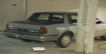 Coche sedán de color gris claro, vista trasera y lateral, estacionado en un interior junto a una columna. Un objeto rectangular blanco se ubica en el suelo