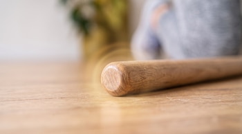 Primer plano del extremo de un palo de madera redondeado sobre un piso, con una persona desenfocada en el fondo.
