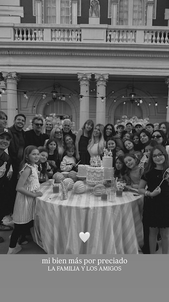 Fotografía en blanco y negro de un grupo numeroso de personas reunidas frente a una mesa con pasteles y un edificio con columnas