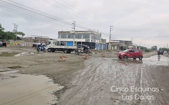 Calle de tierra cubierta de lodo y charcos por la que circulan un automóvil rojo, un mototaxi, un camión y una motocicleta