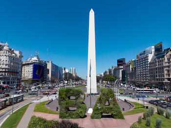 Obelisco de Buenos Aires, un pilar blanco, en medio de una intersección con vehículos, edificios y las letras 'BA' de vegetación. Cielo azul claro