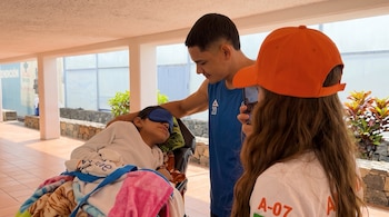 Un niño recostado en una silla lleva un antifaz azul junto a un hombre con camiseta azul y una mujer con gorra naranja.