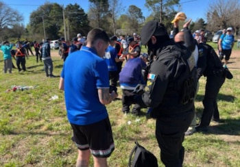 Agentes de seguridad con uniforme negro requisan a varias personas vestidas con camisetas azules en un terreno al aire libre