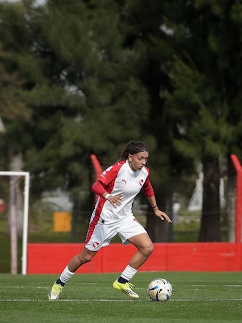 Una jugadora de fútbol con camiseta blanca y roja corre controlando la pelota en un campo verde con árboles de fondo