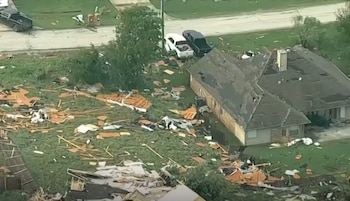 Vista aérea de una casa con tejado dañado y un jardín lleno de escombros de madera y otros materiales. Dos camionetas estacionadas cerca de la carretera