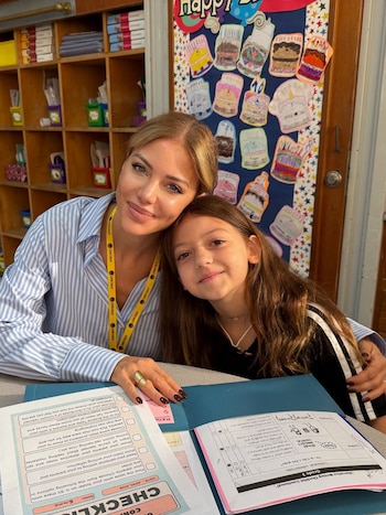 Evangelina Anderson y su hija Emma Demichelis sonríen juntas en un aula, con la niña apoyando su cabeza en la madre, frente a papeles y un estante