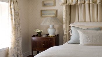 Cama con dosel, almohadas decoradas, mesita de noche de madera, lámpara de mesa, cortinas estampadas, papel tapiz, dos cuadros, jarrón con flores.
