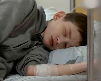 Un joven descansa en una cama de hospital. Viste una sudadera gris, tiene los ojos cerrados y un vendaje con un tubo de vía intravenosa en el brazo